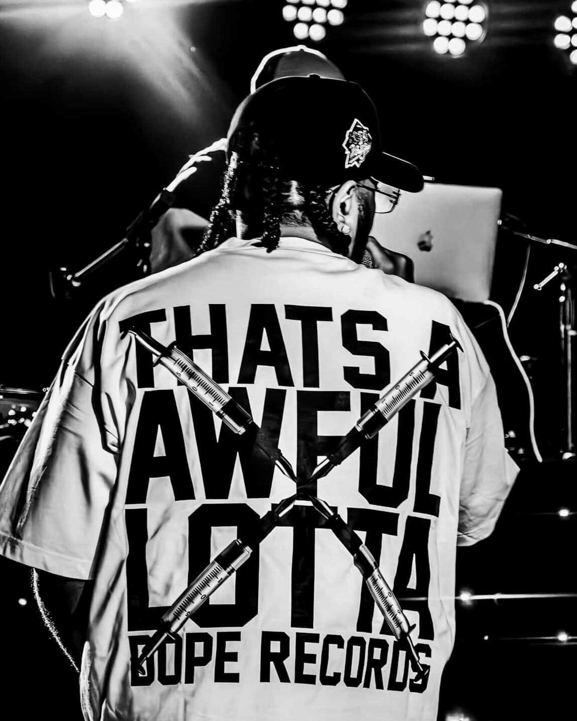 A man in a cap and braids, seen from behind on stage under the lights; the back of his tee reads “Thats a awful lotta Dope Records” with two syringes crossed in an X over the words